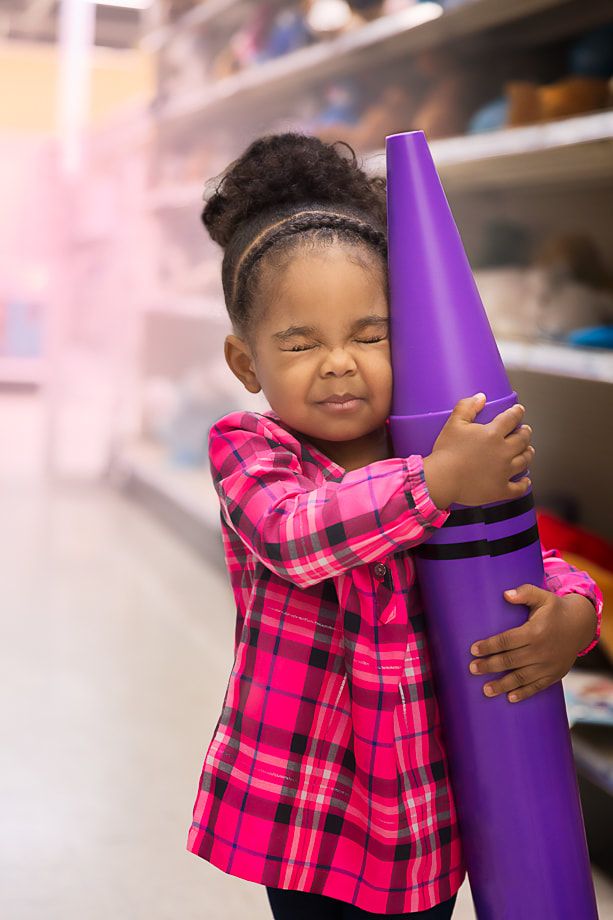 A young girl is posing for child pictures with Woodbridge family photographer Tamieka Smith, hugging a purple crayon. The girl is standing inside Toys R US in Woodbridge, VA, with her eyes squeezed closed and showing her love for the color purple and her happy personality.