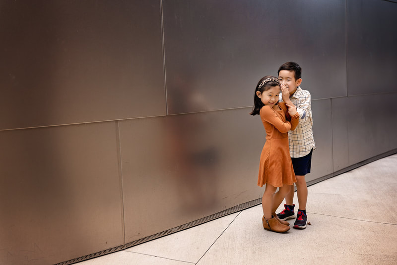 Captured in this heartwarming photograph by Los Angeles Family Photographer Tamieka Smith, two siblings share a special moment at the iconic Disney Concert Hall. The siblings lean in towards each other, giggling and whispering, sharing a secret and strengthening the bond that ties them together. Tamieka's expert use of framing and composition captures the striking architecture of the concert hall, with its curves and lines providing a perfect backdrop to the siblings' intimacy. The lighting accentuates their features and the warmth in their expressions, revealing the beauty and love that connects them. This photograph is a powerful testament to the enduring love and closeness of siblings, and the magic of the world around us that can bring us closer together. It is a moment frozen in time, one that the siblings and their family will treasure forever, a testament to the enduring power of love, joy, and connection.