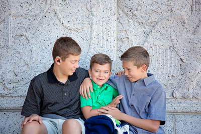 Three brothers enjoy a fun day together, sitting in front of the National Archives Museum in Washington DC, captured by Tamieka Smith Photography. Their laughter fills the air as they pose for the camera, their close bond evident in their relaxed and playful demeanor. The sun shines brightly, illuminating their happy faces and the impressive architecture of the museum behind them.