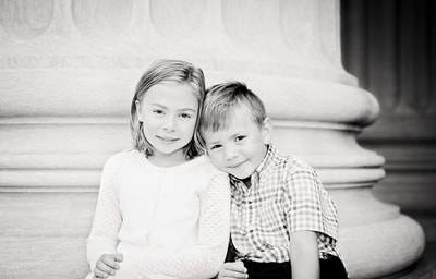 Brother and sister sit on the steps of the National Archives Museum in Washington DC, smiling for a photo taken by Tamieka Smith Photography, a family photographer in Northern Virginia. The sun casts a warm glow on their faces as they lean towards each other, their bond evident in their shared laughter and comfortable closeness.