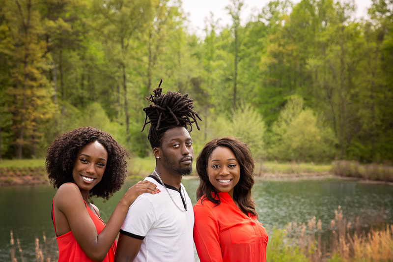 Three siblings are posing for a portrait session with a Northern Virginia family photographer in Woodbridge. The children are standing in a park with a wooded area and pond in the background, wearing coordinating outfits and happily interacting with each other, creating a warm and loving family moment captured in a photograph.