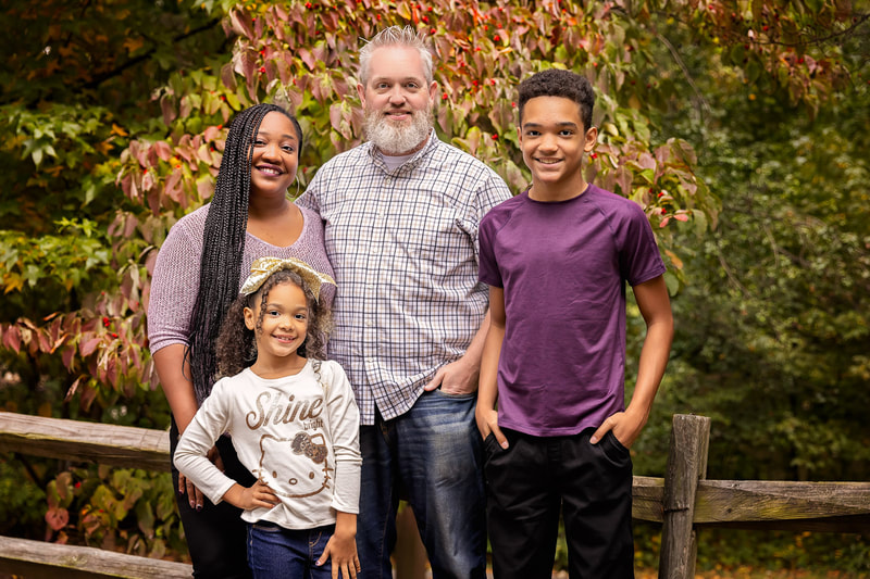 A family portrait session captured in a beautiful outdoor setting in Woodbridge, VA featuring a happy family of four posing together. The image is framed from head to toe, with a natural background of fall trees. This family portrait near me is a cherished keepsake that captures the family's love, joy, and togetherness.